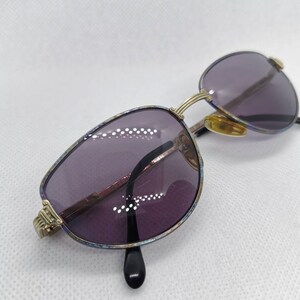 Pode incluir: Um par de óculos de sol com armação dourada, lentes roxas e uma armação com padrão azul e dourado.