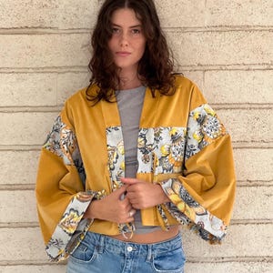 Könnte beinhalten: Senfgelbe Samtjacke mit floral gemusterten Ärmeln und Akzenten. Die Jacke hat eine lockere Passform und einen Bindverschluss. Die Ärmel zeigen ein grau-weißes Blumenmuster mit gelben Akzenten. Die Jacke wird mit einer blauen Jeans kombiniert.