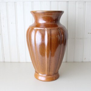 May include: A glossy, medium-brown ceramic vase featuring a wide, flared rim and a body defined by vertical, raised ridges that extend from the neck down to the base, creating a structured, symmetrical silhouette with a smooth, reflective finish.