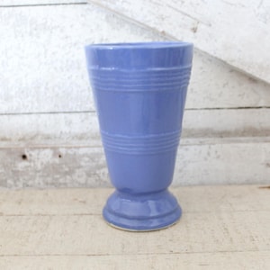 May include: A periwinkle blue ceramic vase with a glossy finish, featuring a flared top opening, a tapered body with two sets of horizontal ribbed bands, and a circular pedestal base that provides stability for the upright vessel structure.