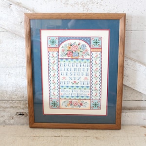 May include: Framed cross-stitch sampler with a floral and alphabet design. The artwork features a blue background, a wooden frame, and the text "Bless This House And Fill All Within."