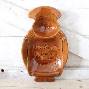 May include: A brown ceramic onion soup dip server in the shape of a chef. The server has a bowl for the dip and a scroll with the recipe. The recipe includes sour cream, onion soup, garlic, and salt.
