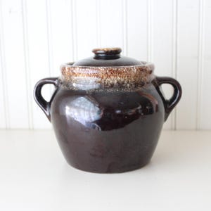 May include: A dark brown ceramic bean pot with two side handles and a matching lid. The upper rim and lid edge feature a textured, mottled cream and tan glaze that contrasts with the smooth, glossy dark brown finish of the main body.