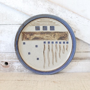 May include: A round ceramic plate with a blue rim and a cream-colored center. The plate features a geometric design with brown and blue accents. The design includes squares, rectangles, and vertical lines. The plate is displayed on a white wooden surface.