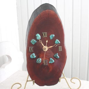 May include: A unique wooden clock with a natural edge, featuring a dark brown finish and turquoise accents. The clock face displays Roman numerals and gold-colored hands. It is mounted on a gold-colored metal stand.