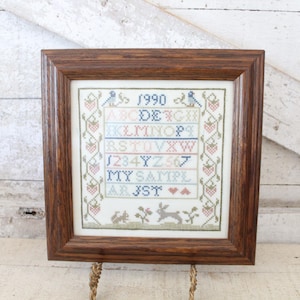 May include: A framed cross-stitch sampler with a brown wooden frame. The sampler features the year "1990" above an alphabet, numbers, and the words "MY SAMPLE" and "AR JST". Decorative elements include birds, strawberries, and a rabbit.