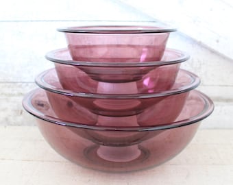 May include: A stack of four vintage glass mixing bowls in varying sizes. The bowls are a translucent purple color with a dark rim. They are arranged on a white wooden surface.