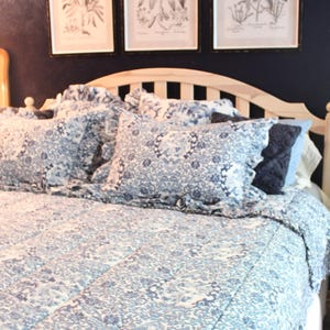 May include: A bed featuring a light-colored wooden headboard and a blue and white patterned quilt with matching ruffled pillow shams. Three framed botanical prints with blue floral illustrations are displayed on the dark wall above the bed.