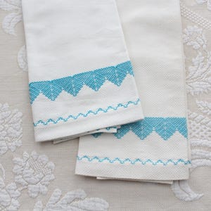 May include: Two white hand towels with a turquoise blue embroidered border. The border features a zigzag and scalloped design. The towels are folded and rest on a cream-colored floral patterned tablecloth.