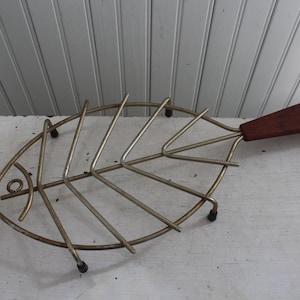 May include: A silver metal fish-shaped hot plate with a wooden handle. The hot plate has a decorative design of fish scales and a small eye.