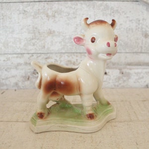May include: A ceramic cow planter with brown and white spots. The cow has a pink nose and pink spots on its cheeks. The planter is standing on a green base.