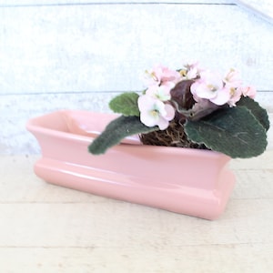 May include: A pink ceramic planter with a long, rectangular shape. It features a delicate, wavy design and holds a small plant with pink flowers and green leaves.