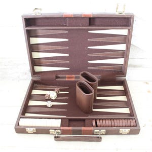 May include: Open brown backgammon set with ivory and brown playing pieces. The set includes dice, dice cups, and playing stones. The case is open, revealing the playing surface with triangular points. The set is on a white surface.