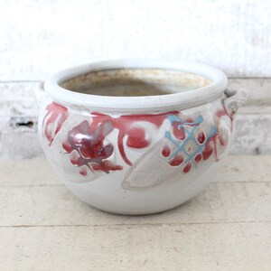 May include: A round, white ceramic planter with a wide rim and two small handles. The planter is decorated with red and blue floral designs. The interior of the planter is a light brown color.