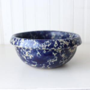 May include: A round ceramic mixing bowl featuring a deep blue base color with a white sponge-painted or mottled pattern throughout the exterior and interior rim. The bowl has a thick, rounded top edge and a smooth, curved basin shape.