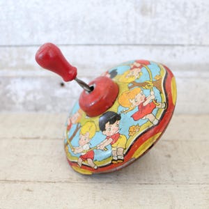 May include: Vintage tin toy top with a red handle and a colorful design featuring children. The top has a red and yellow rim, with a blue and yellow background. The children are wearing red and yellow clothing.