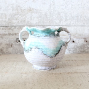 May include: A small, white ceramic vase with two handles and a light blue and white glaze. The vase has a scalloped rim and a textured surface.
