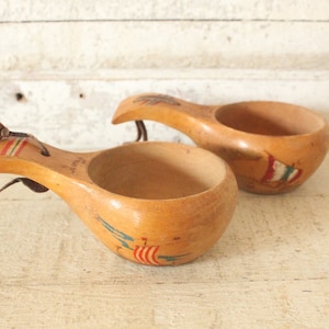 May include: Two wooden cups with leather handles and painted designs of sailing ships. The cups are light brown with red, blue, and green accents.