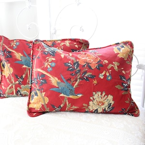 May include: Two decorative throw pillows with a vibrant red background and a floral and bird pattern. The pillows feature a mix of colors, including blue, yellow, and orange, and are placed on a white bed.