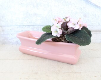 May include: A pink ceramic planter with a long, rectangular shape. It features a delicate, wavy design and holds a small plant with pink flowers and green leaves.