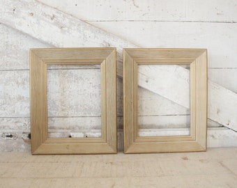 May include: Two gold-toned picture frames with a simple, rectangular design. The frames are made of wood and have a light, natural finish. They are sitting on a white wooden surface.