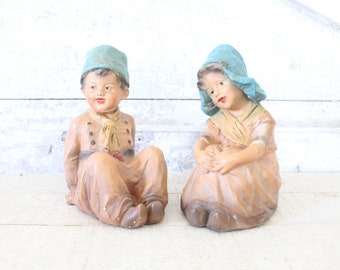 May include: Two vintage ceramic figurines of children wearing blue hats. The boy is wearing a brown and tan outfit and the girl is wearing a pink and tan dress. The figurines are sitting on a white wooden surface.
