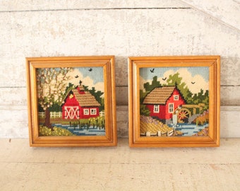 May include: Two framed needlepoint pictures of red houses with a country scene. The picture on the left has a house with a white fence and a tree with white blossoms. The picture on the right has a house with a water wheel and a river.