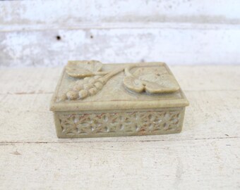 May include: A small, square, stone box with an intricate floral design carved into the top. The box is made of a light brown stone and has a detailed, carved pattern around the edges.