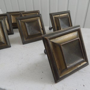 May include: A set of ten antique brass cabinet knobs with a square shape and a raised, beveled center. The knobs have a distressed, aged finish.