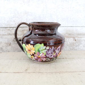 May include: A brown ceramic pitcher with a floral design. The pitcher has a wide base and a rounded body. The flowers are painted in shades of yellow, purple, and green.