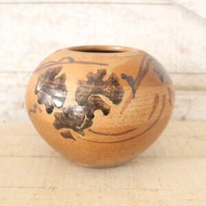 May include: A round, brown ceramic vase with a black and white floral design. The vase has a glossy finish and is decorated with stylized leaves and branches.