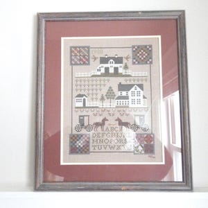 May include: Framed cross-stitch artwork featuring a rural scene with houses, trees, and horse-drawn carriages. The piece includes the year "1908" and an alphabet. The artwork is framed in a gray wooden frame with a burgundy mat.