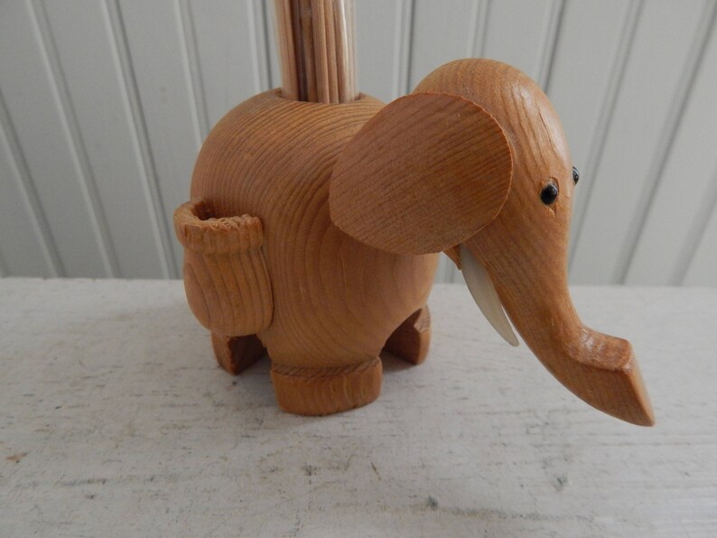 Puede incluir: Porta palillos de dientes de madera con forma de elefante con una ranura para los palillos en el lateral. El elefante est&aacute; hecho de madera marr&oacute;n claro y tiene ojos negros.