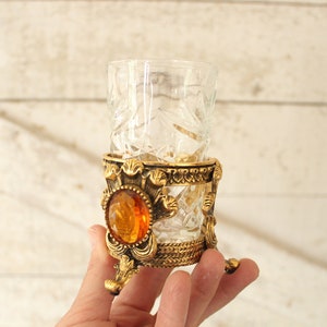 May include: A clear glass shot glass with a cut glass design sits on a gold metal stand with an amber jewel accent.