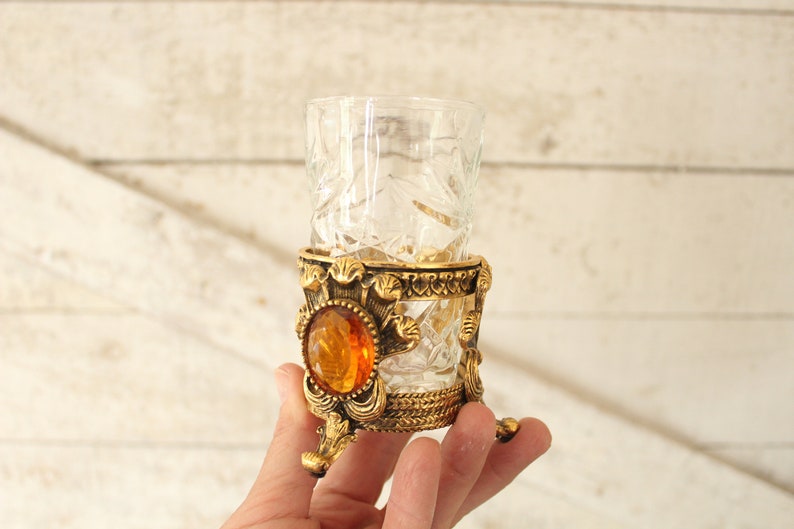 May include: A clear glass shot glass with a cut glass design sits on a gold metal stand with an amber jewel accent.