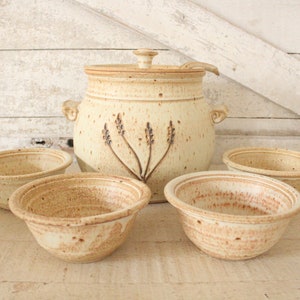 May include: A set of five ceramic bowls, including a large serving bowl with a lid and a ladle. The bowls are all a light beige color with brown speckles and a design of lavender flowers.