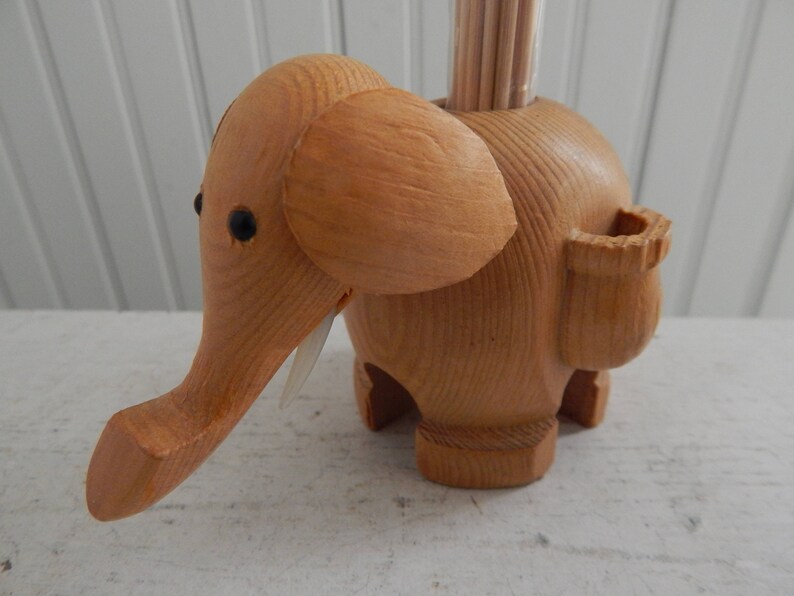 Puede incluir: Porta palillos de dientes de madera con forma de elefante con una ranura para palillos en el lateral. El elefante es marr&oacute;n y tiene ojos negros.