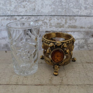 May include: A clear glass tumbler with a cut-glass design sits next to a gold-tone decorative stand with a round top and three legs. The stand has a large amber-colored jewel in the center.