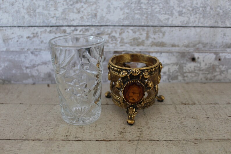 May include: A clear glass tumbler with a cut-glass design sits next to a gold-tone decorative stand with a round top and three legs. The stand has a large amber-colored jewel in the center.
