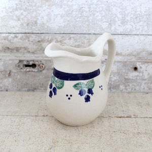 May include: A white ceramic creamer with a blue and green floral design. The creamer has a handle and a spout.
