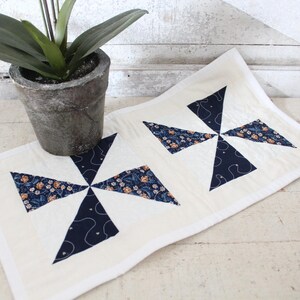 May include: Cream-colored quilted table runner with two pinwheel designs. Each pinwheel features navy blue and floral patterned fabric. A potted plant sits to the left of the runner. The runner is approximately 12 inches wide.