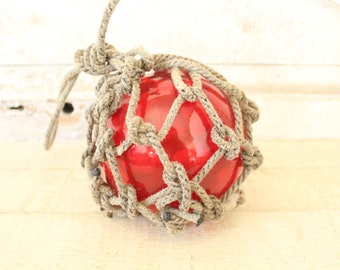 May include: A red glass buoy encased in a thick, natural rope net. The buoy is a traditional nautical decoration.
