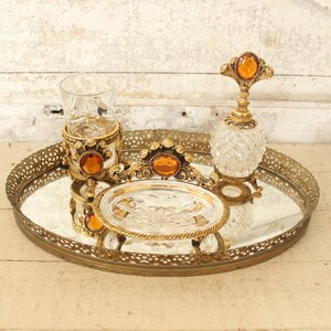 May include: A gold-toned mirrored tray with an ornate design, featuring a glass perfume bottle with a gold top and a glass cup with a gold base. The tray has a decorative design with a gold finish.