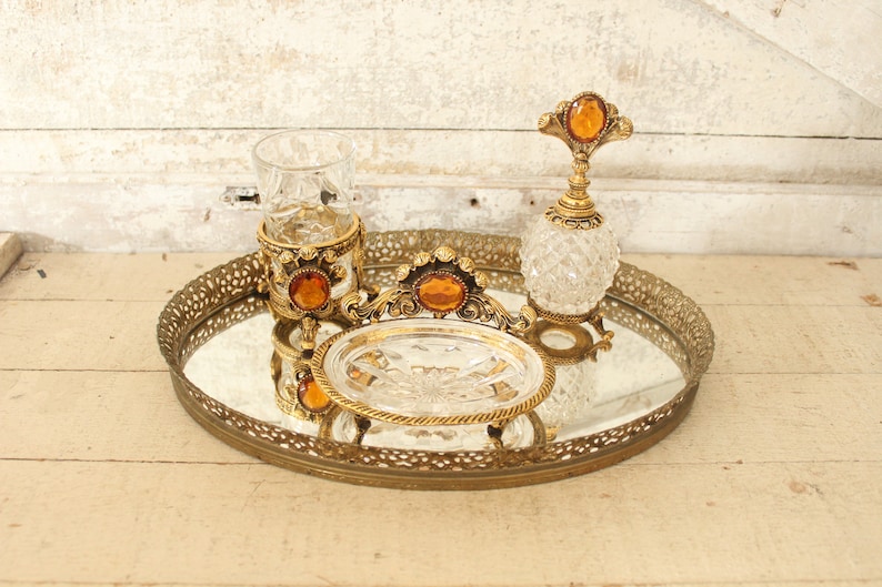 May include: A gold-toned mirrored tray with an ornate design, featuring a glass perfume bottle with a gold top and a glass cup with a gold base. The tray has a decorative design with a gold finish.