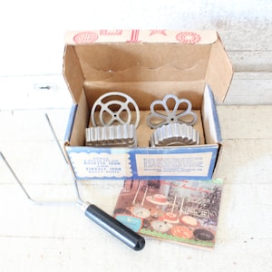 May include: A vintage set of four metal donut molds in a cardboard box with a booklet of recipes. The molds are in the shapes of a flower and a circle. The box has the words "Donut Iron" and "Variety Cakes" printed on it.