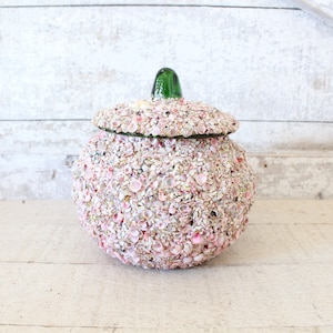 May include: A round, white jar with a green glass lid, completely covered in small pink and white seashells.