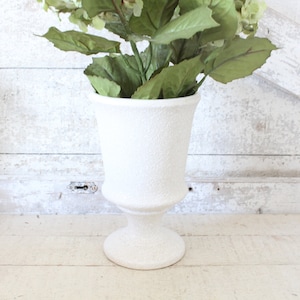 May include: White ceramic vase with a textured surface, featuring a classic urn shape with a pedestal base. The vase is filled with green leafy stems and white flowers.