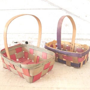 May include: Two woven baskets with arched handles. One basket features red and tan woven strips, while the other has purple and tan strips. Both baskets are rectangular with a wooden handle.