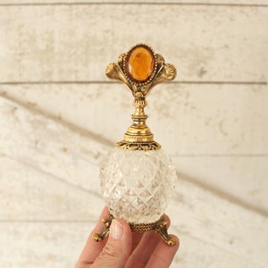 May include: A clear glass bottle with a gold metal top and a decorative amber stone. The bottle has a faceted design and sits on three gold metal legs.