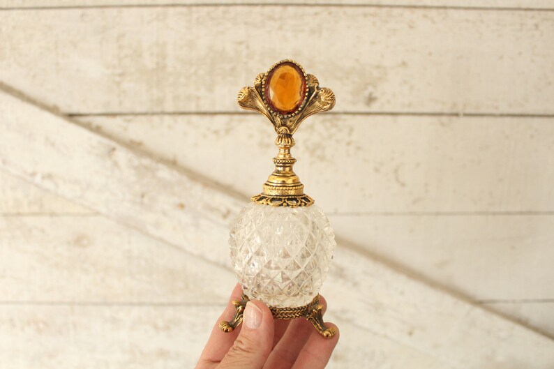 May include: A clear glass bottle with a gold metal top and a decorative amber stone. The bottle has a faceted design and sits on three gold metal legs.